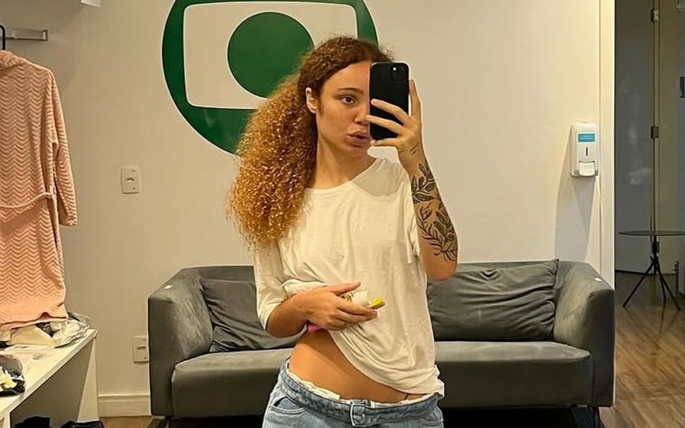 Leticia Vieira tira foto no espelho em camarim; na parede ao fundo há o logo da Globo