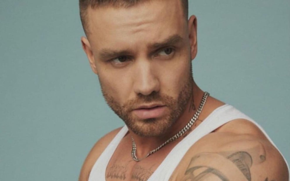 Liam Payne está com o corpo de lado, veste uma regata branca, e olha para a esquerda