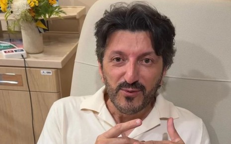 homem moreno veste uma camisa branca e fala com a câmera deitado em cama de hospital