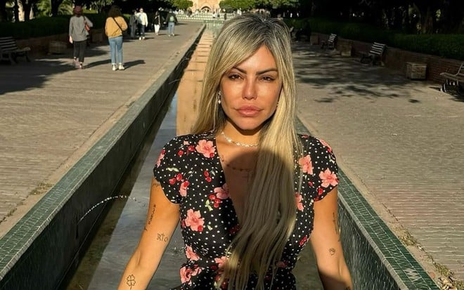 Liziane Gutierrez está com um vestido florido, séria e sem maquiagem; ela encara a câmera