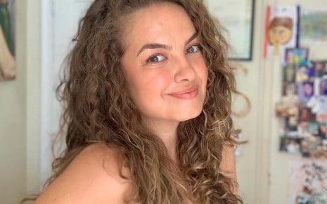 A atriz Lua Blanco surge em primeiro plano, com cabelos castanhos volumosos e encaracolados caindo sobre os ombros, inclinando ligeiramente a cabeça e sorrindo de forma suave para a câmara num ambiente interior.