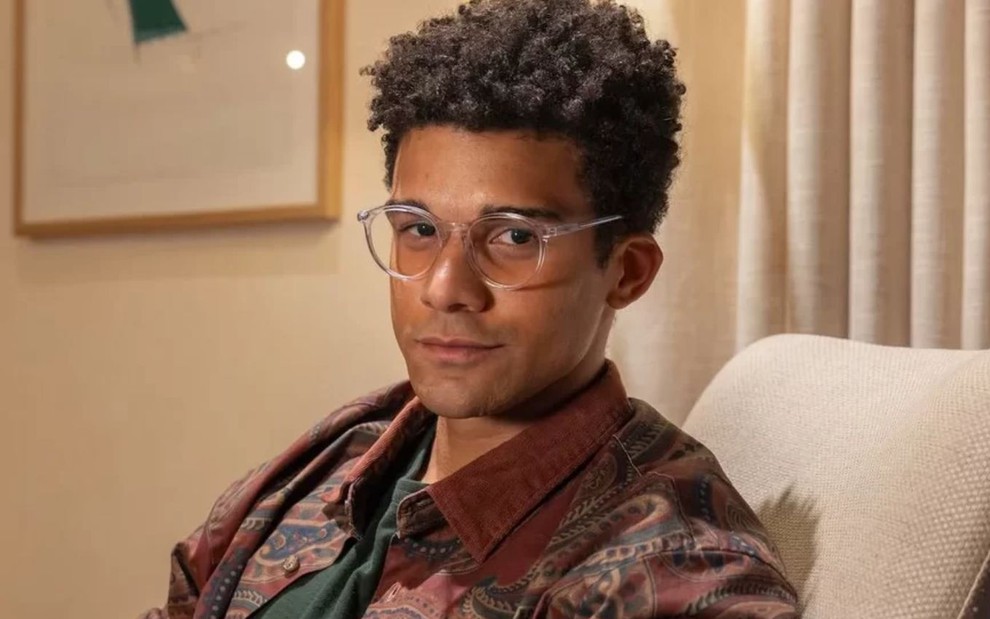 Lucas Leto como Sardinha em Vale Tudo; ele está sentado em uma poltrona branca, usando camisa marrom e óculos