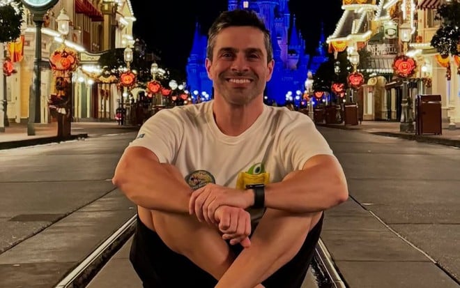 Luciano Amaral sentado em frente ao castelo da Cinderela na Disney; em cenário noturno, o castelo está iluminado de azul