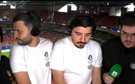 Três homens em transmissão esportiva. Dois vestem camiseta branca e fones; o da direita, de óculos, segura microfone verde. Fundo de estádio