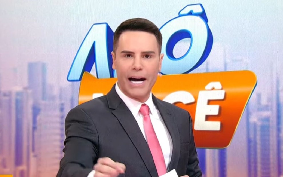 Apresentador gesticula com expressão séria em frente ao letreiro colorido do programa “Alô, Você"