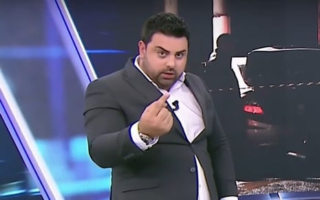 Homem de terno faz gesto com os dedos em frente a telão, com expressão séria e olhar direto para a câmera.