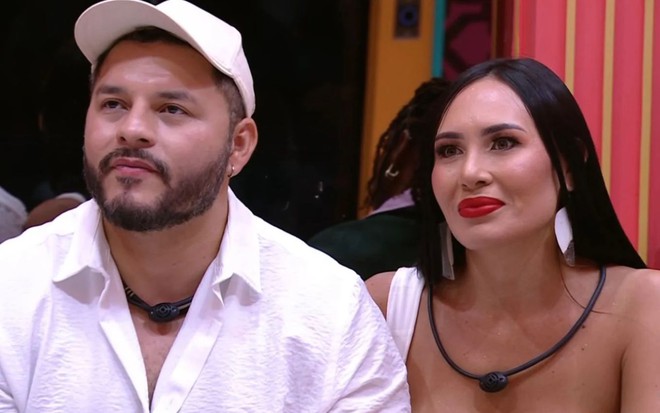 Marcelo e Arleane sentados no sofá do BBB 25, no dia de sua eliminação; ele usa camisa branca e boné branco, e ela usa regata branca