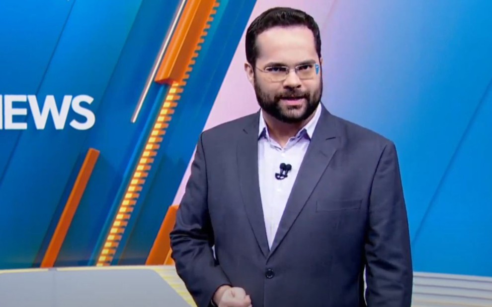 Apresentador em estúdio de TV com fundo azul e detalhes laranja. Usa terno cinza, camisa branca e óculos, com microfone de lapela.