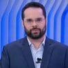 O jornalista Marcelo Casagrande usa terno, está com expressão séria e parado em frente a telão com logo do SBT News