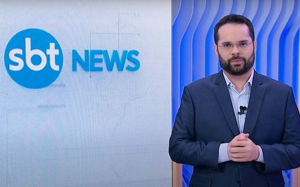 O jornalista Marcelo Casagrande usa terno, está com expressão séria e parado em frente a telão com logo do SBT News