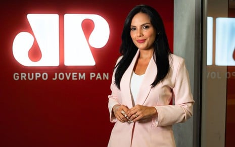 Márcia Dantas posando com terno bege em frente ao logo da Jovem Pan