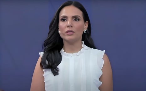 A mulher da imagem parece estar apresentando ou discursando, com expressão séria e cabelos escuros, usando uma blusa branca elegante.