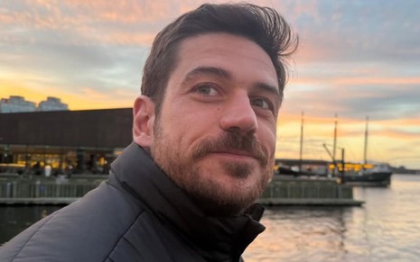 Marco Pigossi tira uma selfie; ele usa casaco preto, enquanto há um rio ao fundo