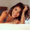 Maria Carol Rebello está deitada de bruços em cama; ela apoia a cabeça com a mão e sorri