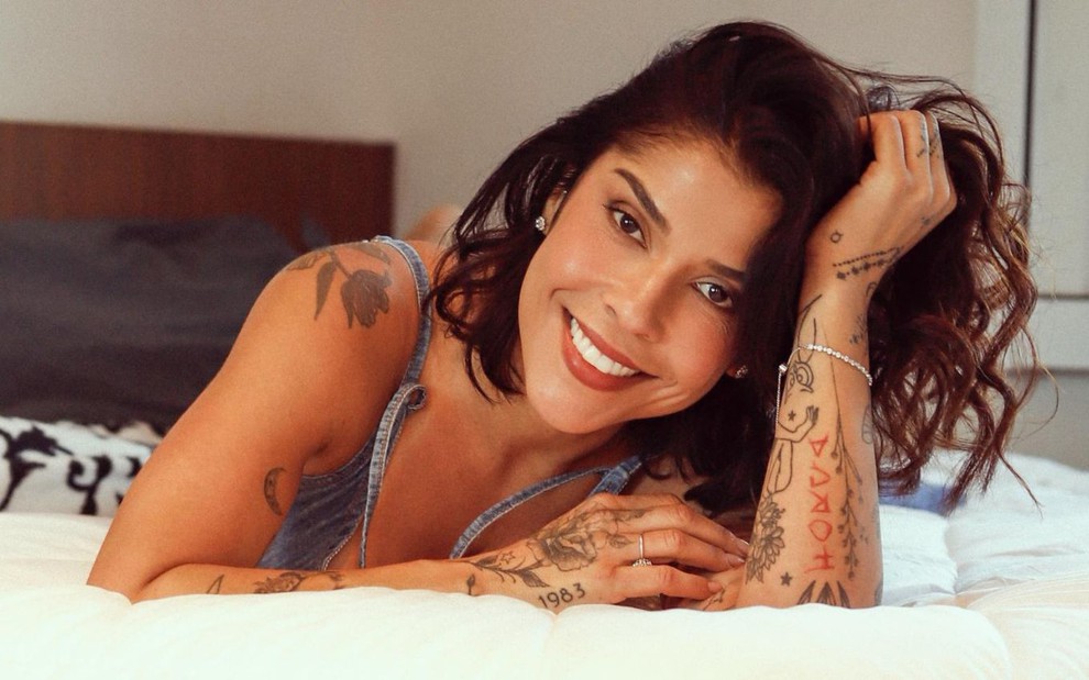 Maria Carol Rebello está deitada de bruços em cama; ela apoia a cabeça com a mão e sorri