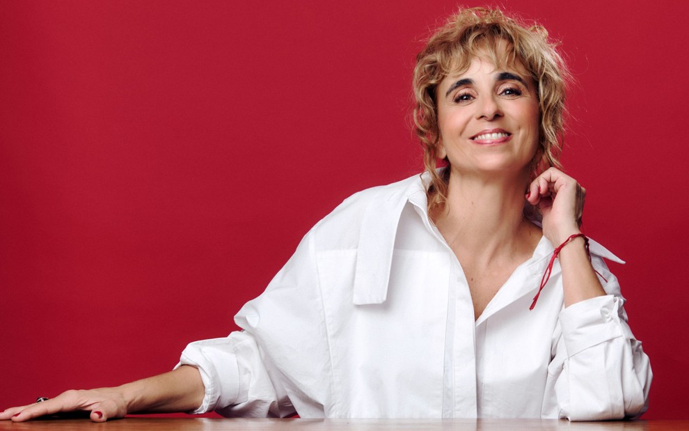 Com camisa social branca, Maria de Medicis posa sorridente em um fundo vermelho