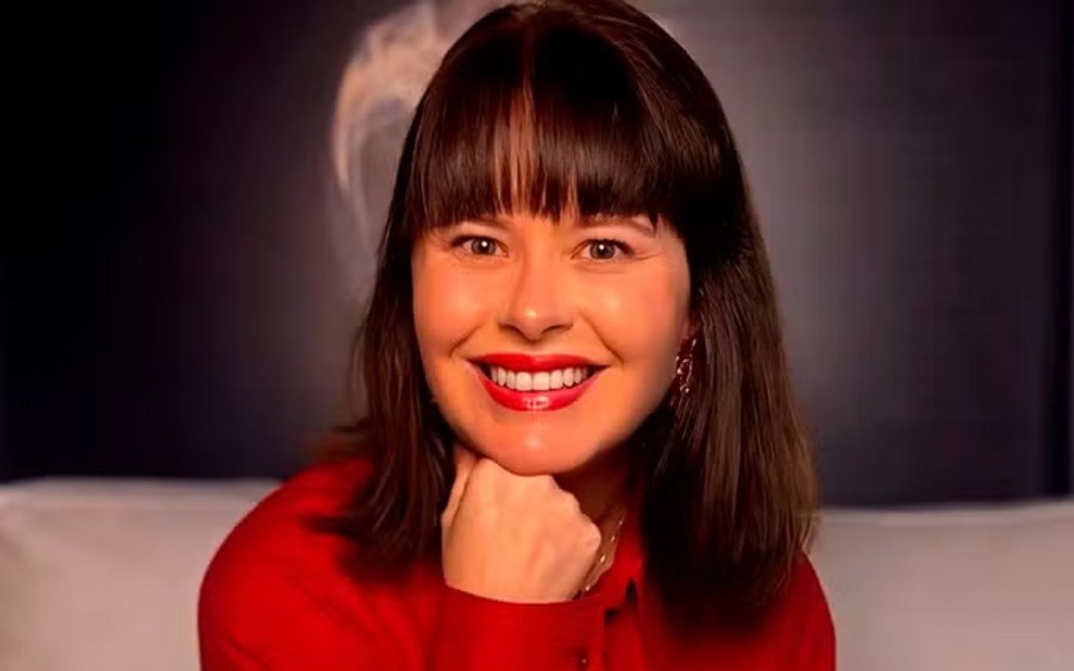 Mariana Bridi está sorridente, com uma das mãos apoiadas no rosto