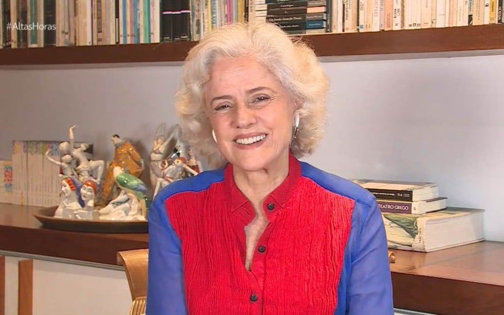 A atriz Marieta Severo sorrindo, com camisa vermelha e azul, em sala de sua casa