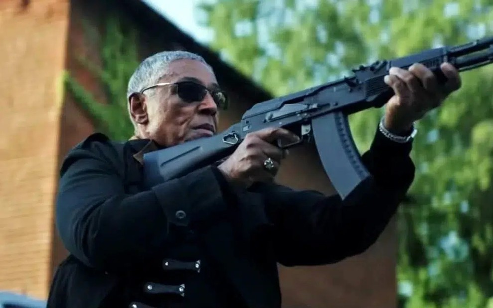 Com roupas e óculos escuros, Giancarlo Esposito aponta uma arma para o alto