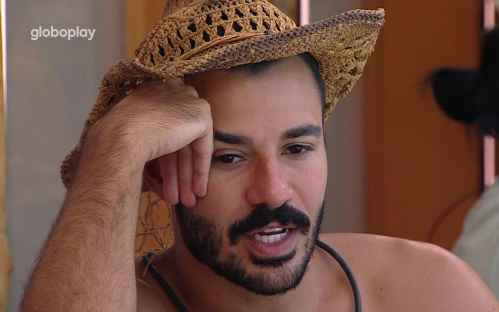 Mateus Pires está com um chapéu de palha, uma das mãos apoiadas no rosto e conversa no BBB 25