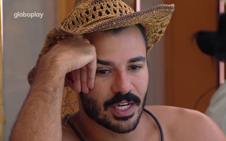 Mateus Pires está com um chapéu de palha, uma das mãos apoiadas no rosto e conversa no BBB 25