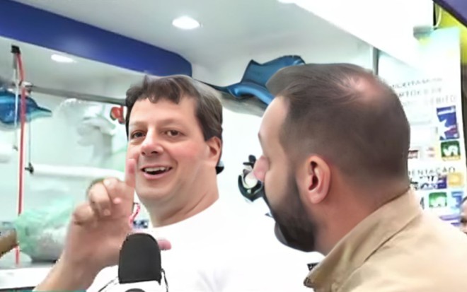 Um homem faz um L com as mãos enquanto é entrevistado por um repórter da Globo
