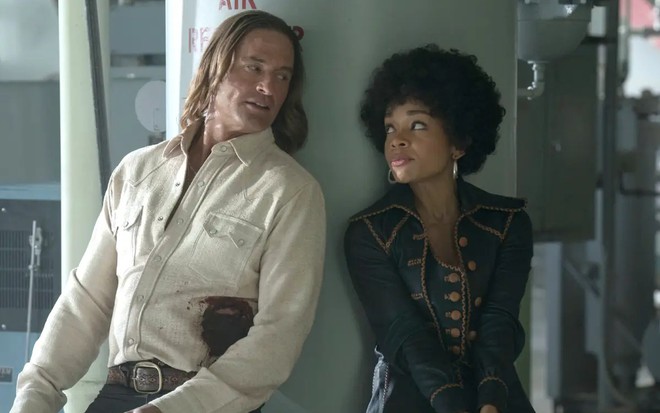 Josh Holloway e Rachel Hilson estão encostados em um pilar; eles usam trajes típicos dos anos 1970, e ela tem cabelo black power
