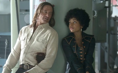 Josh Holloway e Rachel Hilson estão encostados em um pilar; eles usam trajes típicos dos anos 1970, e ela tem cabelo black power