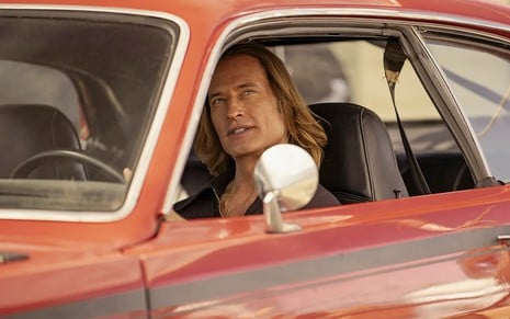 Com cabelos compridos, Josh Holloway está dentro de um carro vermelho e tem uma expressão sacana
