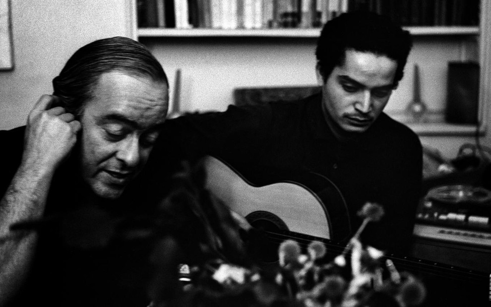 Em foto preto e branca, dois homens estão em uma sala; o primeiro tapa um ouvido, enquanto o segundo toca violão