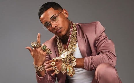 MC Poze do Rodo posando com um terno rosa brilhante, exibindo colares e anéis de ouro