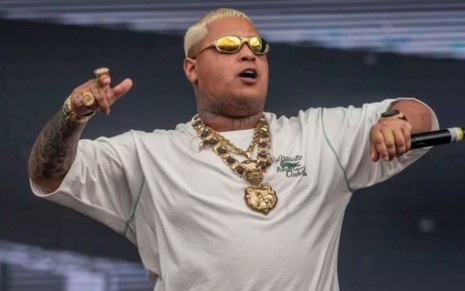 MC Ryan SP está trajado de óculos de sol espelhado e corrente, e segura um microfone