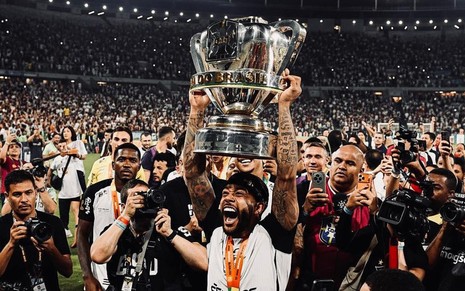 jogador de futebol negro levanta troféu em estádio; ele é cercado de fotógrafos