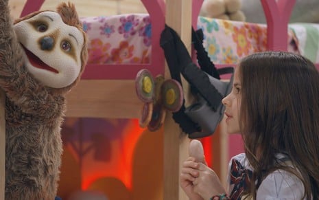 Anna (Mel Summers) segura pedra na mãe e olha para bicho preguiça de pelúcia em cena da novela A Caverna Encantada