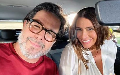 Murilo Benício e Giovanna Antonelli sorridentes, lado a lado, dentro de um carro; ele usa camiseta vermelha e óculos, e ela usa uma blusa branca