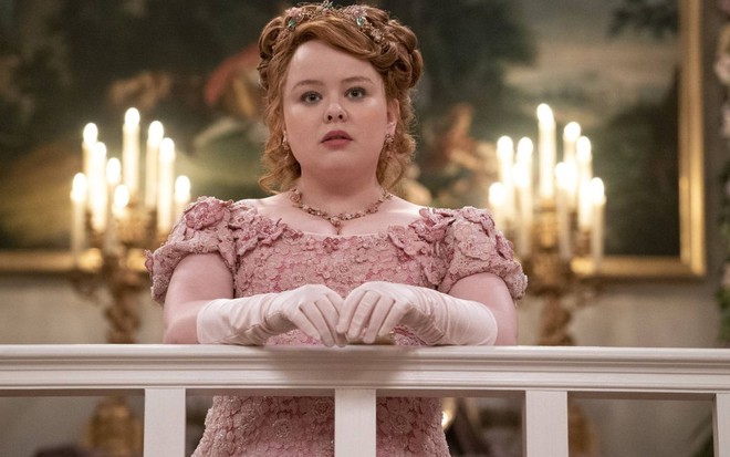 Com vestido cor-de-rosa, Nicola Coughlan está no alto de uma sacada, apoiada no corrimão, com expressão séria