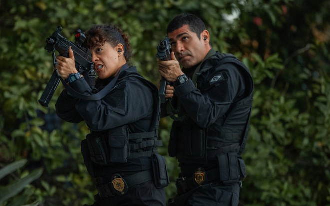 Maeve Jinkings e Rômulo Braga estão com roupas de policiais e empunham metralhadoras em cena de DNA do Crime