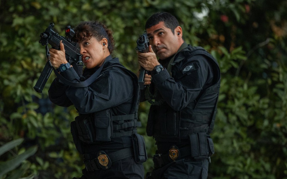Maeve Jinkings e Rômulo Braga estão com roupas de policiais e empunham metralhadoras em cena de DNA do Crime