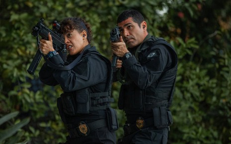 Maeve Jinkings e Rômulo Braga estão com roupas de policiais e empunham metralhadoras em cena de DNA do Crime