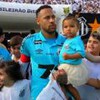 O jogador Neymar, com um uniforme azul do Santos, com a filha Mavie no colo e cercado de crianças na entrada de um campo de futebol