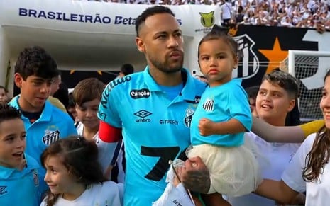 O jogador Neymar, com um uniforme azul do Santos, com a filha Mavie no colo e cercado de crianças na entrada de um campo de futebol