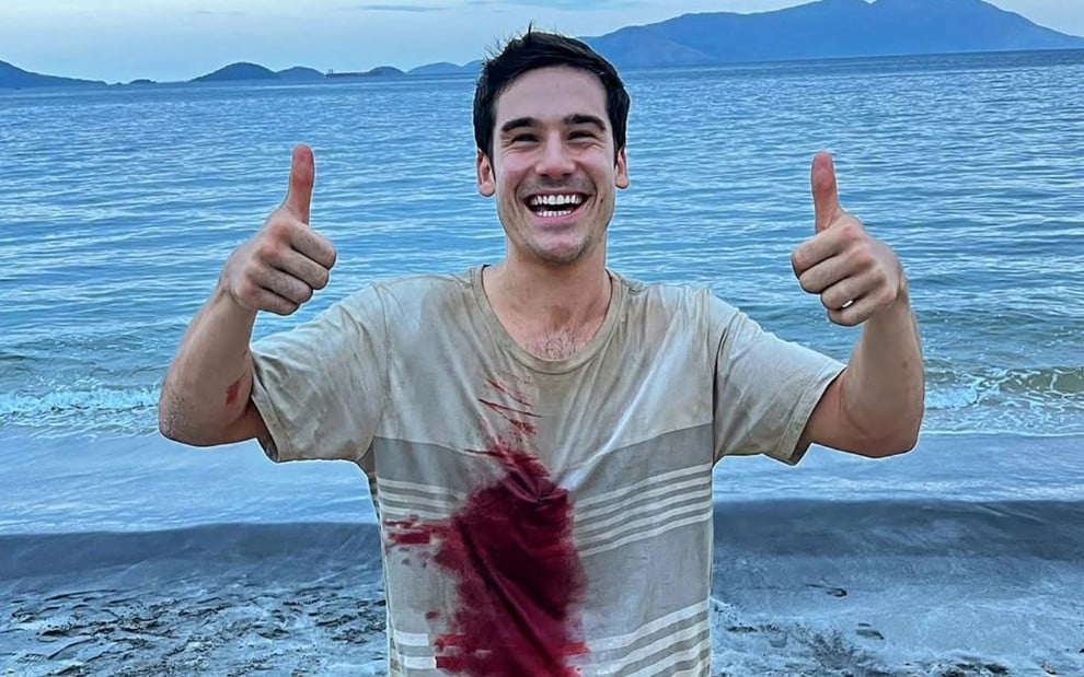 Nicolas Prattes faz sinal de joinha com as duas mãos, sorri e está com mancha de sangue na camiseta; ao fundo é possível ver o mar