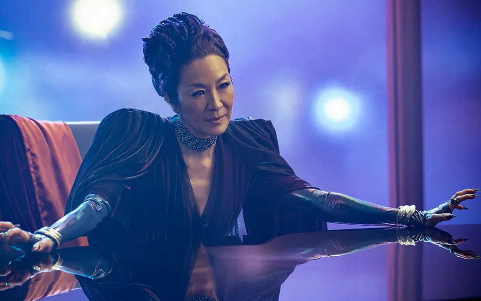 Michelle Yeoh está sentada à mesa, com os braços abertos e um visual futurista