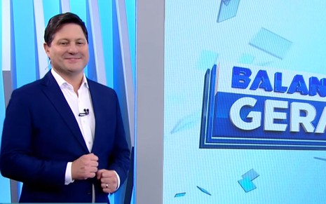 O apresentador Eleandro Passaia, de terno azul-escuro e camisa branca sem gravata, sorri no estúdio ao lado do telão do programa Balanço Geral.