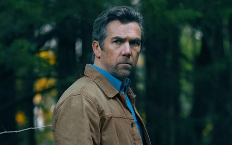 homem branco faz cara de bravo para a câmera em meio a uma floresta; ele veste uma jaqueta marrom e camisa cinza