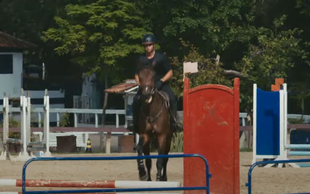 Um homem está montado em um cavalo, em um haras, no trailer de Vale Tudo