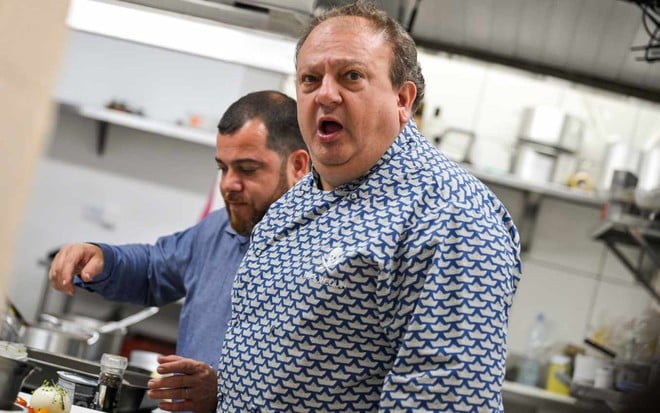 Erick Jacquin durante gravação da nova temporada do Pesadelo na Cozinha, com expressão de irritação em cozinha de restaurante, com outro cozinheiro atrás