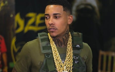 Homem com colete tático verde, várias correntes de ouro no pescoço e tatuagem no pescoço, olhando sério para o lado.