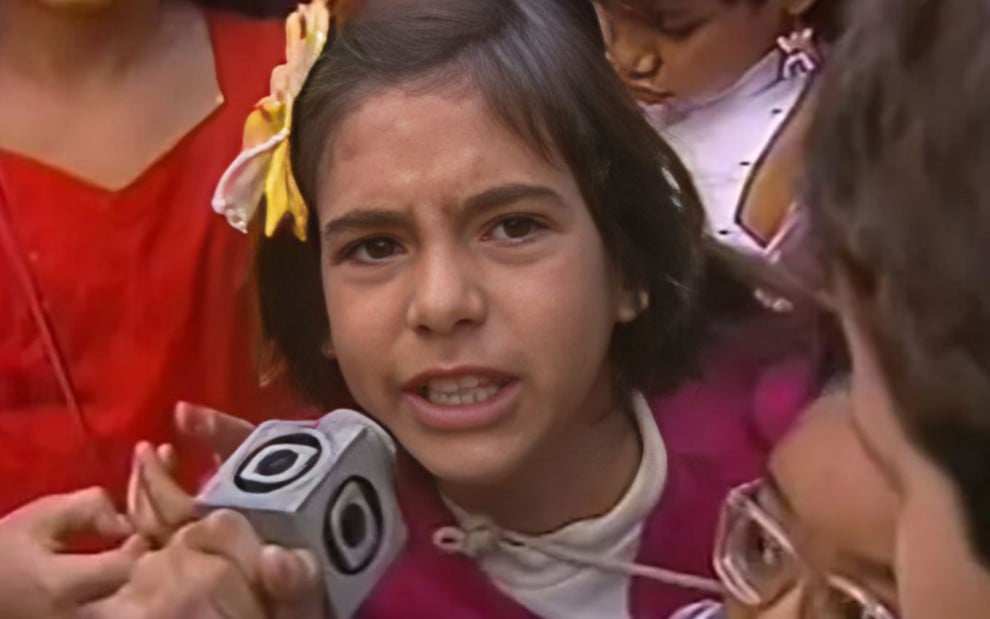 A imagem mostra uma criança segurando um microfone da Globo em um ambiente com outras crianças ao fundo. A menina parece estar falando com alguém, provavelmente respondendo a uma pergunta de um repórter. Ela está usando uma roupa com detalhes coloridos