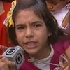 A imagem mostra uma criança segurando um microfone da Globo em um ambiente com outras crianças ao fundo. A menina parece estar falando com alguém, provavelmente respondendo a uma pergunta de um repórter. Ela está usando uma roupa com detalhes coloridos